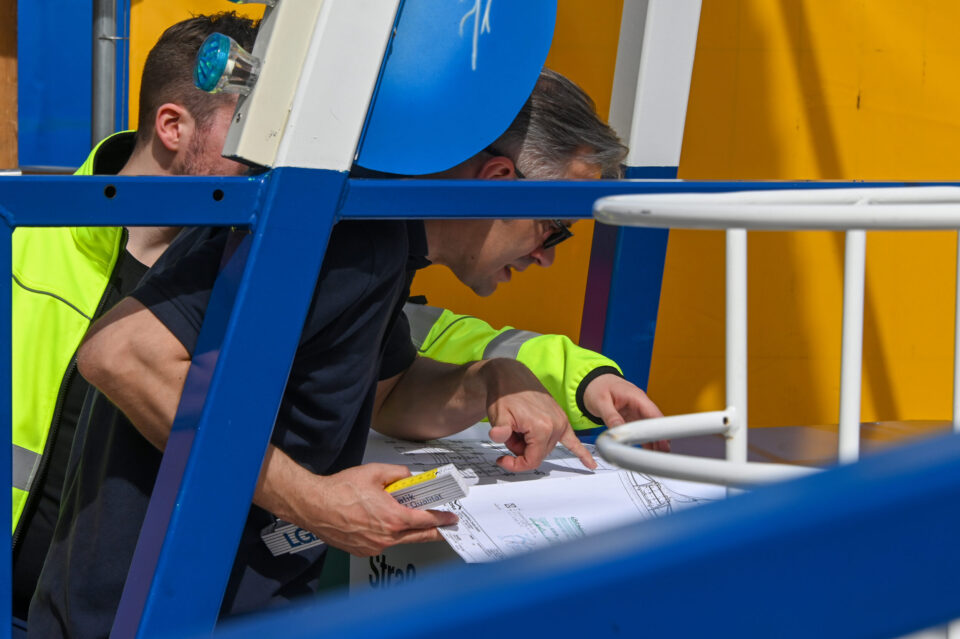 Zwei Personen betrachten technische Zeichnungen oder Baupläne, eine davon trägt eine Warnschutzjacke, während die andere auf die Dokumente zeigt.