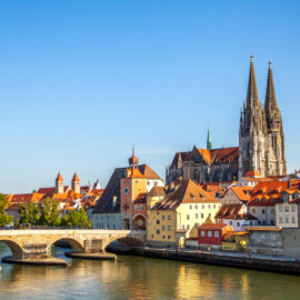Panoramablick auf Regensburg mit dem markanten Dom mit zwei Türmen, einer historischen Steinbrücke über die Donau im Vordergrund und bunten Gebäuden mit roten Dächern entlang des Flussufers bei klarem Himmel.