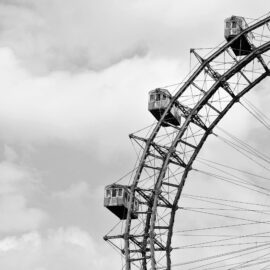 Schwarz-weiß-Fotografie eines historischen Riesenrads, bei der der obere Teil mit mehreren Gondeln und dem filigranen Metallgerüst vor einem wolkigen Himmel zu sehen ist.