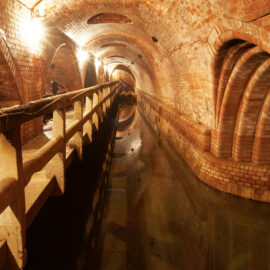 Das Bild zeigt einen unterirdischen Tunnel, Kanal- und Abwasseranlage mit gemauerten Wänden und einem gewölbten Deckengewölbe. Auf der linken Seite verläuft ein schmaler Gehweg, der durch ein Betongeländer vom Wasserkanal getrennt ist. Der Tunnel ist durch mehrere helle Wandlampen beleuchtet, deren Licht sich auf der Wasseroberfläche spiegelt.