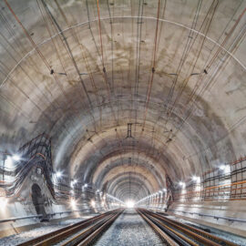 Das Bild zeigt das Innere eines großen, gut beleuchteten Tunnels mit zwei Gleisen. Entlang der Tunnelwände verlaufen verschiedene Kabel und Rohre, und auf beiden Seiten sind regelmäßig Lampen angebracht. Der Blick führt in Richtung eines hellen Lichtpunkts am Ende des Tunnels.