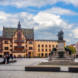 Stadtplatz in Schweinfurt mit einem zentralen Denkmal auf einem Sockel und zwei sitzenden Figuren, im Hintergrund ein reich verziertes Gebäude mit steilem Dach, vermutlich das Rathaus; umgeben von bunten Gebäuden, Kopfsteinpflaster und Menschen bei teils bewölktem Himmel.