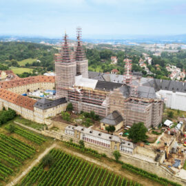 Luftaufnahme der ehemaligen Abteikirche St. Michael in Bamberg mit sichtbaren Restaurierungsarbeiten an den Türmen; umgeben von Gebäuden mit roten Dächern, einem großen Innenhof sowie Weinbergen und Gärten im Vordergrund.