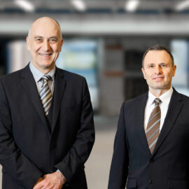 Zwei Personen in dunklen Anzügen mit weißen Hemden und gestreiften Krawatten stehen in einem Foyer. Der Hintergund ist stark verschwommen.