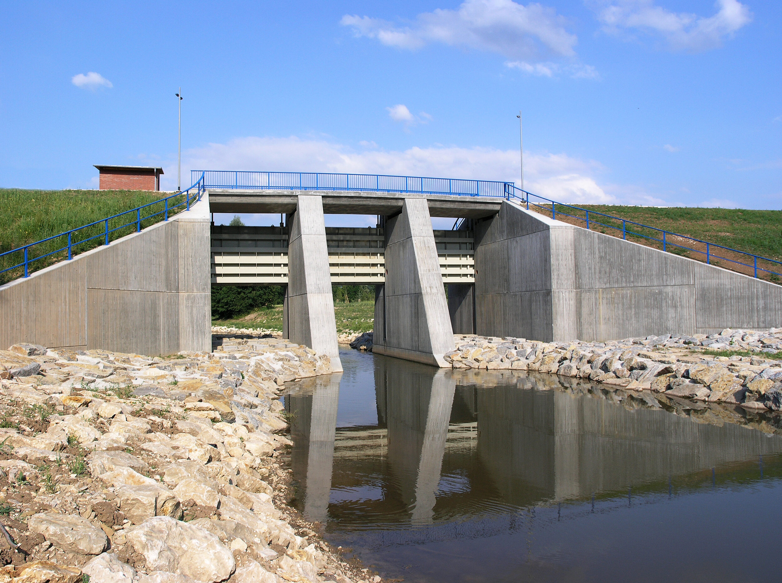 Das Bild zeigt eine Betonbrücke über einem kleinen Gewässer. Die Brücke hat drei große rechteckige Wasserdurchlässe, die von massiven Betonpfeilern getragen werden. Sie dienen als Staudamm. Entlang der Brücke verläuft ein blaues Metallgeländer, die Ufer sind mit Steinen gegen Erosion gesichert.