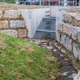 Das Bild zeigt eine Hochwasserschutzanlage mit einem Betonkanal, an dessen Eingang ein Metallgitter angebracht ist, um Treibgut fernzuhalten. Die Umgebung ist mit großen Steinblöcken gesichert, um Erosion zu verhindern und die Ufer zu stabilisieren. Auf der Anlage befindet sich ein Metallgeländer als Sicherheitsmaßnahme.