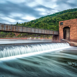 Das Bild zeigt einen Damm, über den Wasser hinabfließt und dabei einen Wasserfall bildet. Über dem Damm verläuft eine Brückenkonstruktion, die zwei große Backsteintürme auf beiden Seiten des Wasserlaufs verbindet. Im Hintergrund ist ein bewaldeter Hügel mit dichter Vegetation zu sehen, darüber ein wolkiger Himmel mit Lichtstreifen.
