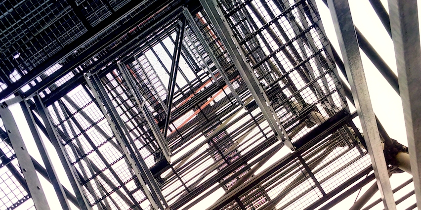 Das Bild zeigt eine Metallkonstruktion, ein Treppenaufgang aus der Froschperspektive. Mehrere Ebenen mit gitterartigen Plattformen erzeugen ein geometrisches Muster, das sich zur Bildmitte hin verjüngt.