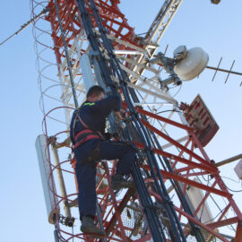 Das Bild zeigt einen Arbeiter, der einen hohen Kommunikationsturm erklimmt. Der Turm besteht aus rot-weiß gestrichenen Metallstrukturen und ist mit verschiedenen Antennen und Satellitenschüsseln ausgestattet. Der Arbeiter trägt Sicherheitsausrüstung einschließlich eines Gurts und führt Wartungsarbeiten durch.