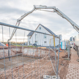 Das Bild zeigt eine Baustelle mit einem großen Betonpumpenfahrzeug, dessen Ausleger über ein abgesperrtes Fundamentbereich reicht. Im Hintergrund steht ein modernes weißes Haus mit dunklem Dach. Auf der Baustelle sind verschiedene Baumaterialien und Geräte verteilt, der Himmel ist teilweise bewölkt.