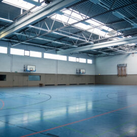 Bauwerksprüfung nach Richtlinie VDI 6200 Standsicherheit Halle LGA in Bayern. Das Bild zeigt eine leere Turnhalle mit einem blauen Boden, der mit Linien für verschiedene Sportarten markiert ist. Die Halle hat eine hohe Decke mit Metallträgern und Oberlichtern, durch die Tageslicht einfällt. An den Wänden befinden sich zwei Basketballkörbe sowie ein zusammengeklappter Kletterrahmen auf der rechten Seite.