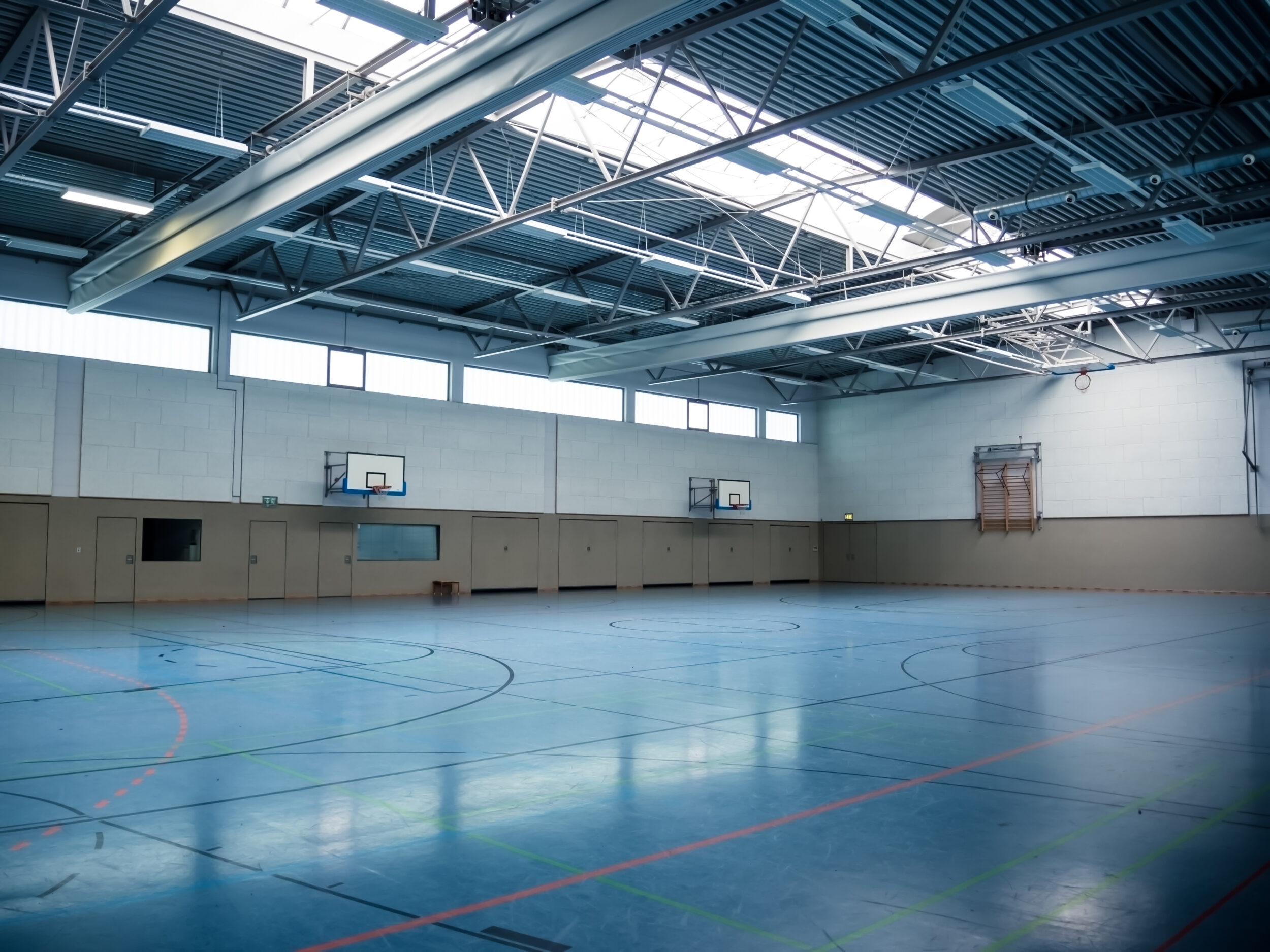 Bauwerksprüfung nach Richtlinie VDI 6200 Standsicherheit Halle LGA in Bayern. Das Bild zeigt eine leere Turnhalle mit einem blauen Boden, der mit Linien für verschiedene Sportarten markiert ist. Die Halle hat eine hohe Decke mit Metallträgern und Oberlichtern, durch die Tageslicht einfällt. An den Wänden befinden sich zwei Basketballkörbe sowie ein zusammengeklappter Kletterrahmen auf der rechten Seite.