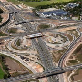 Das Bild zeigt eine Großbaustelle am Autobahnkreuz A3 mit mehreren Fahrspuren und Rampen. In der Mitte des Kreuzes sind Bauarbeiten im Gange, sichtbar durch Baumaschinen und Geräte. Umgeben ist das Areal von Feldern, Wohngebieten und Industrieanlagen.