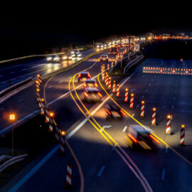 Das Bild zeigt eine nächtliche Szene auf einer mehrspurigen Autobahn mit mehreren fahrenden Autos. Durch die Langzeitbelichtung entstehen Lichtspuren von Scheinwerfern und Rücklichtern. Zahlreiche Verkehrsleitkegel und Absperrungen mit reflektierenden Streifen deuten auf Bauarbeiten hin.