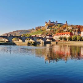 Ansicht von Würzburg mit einem historischen Steinbogenbrücke über einen ruhigen Fluss im Vordergrund, dahinter Gebäude mit roten Dächern entlang des Ufers und eine große Festung auf einem Hügel, umgeben von herbstlich gefärbten Bäumen unter blauem Himmel.